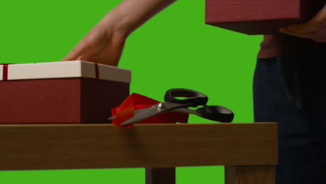 Close-Up-Of-Man-Picking-Up-Gift-Wrapped-Presents-In-Boxes-Decorated-With-Ribbon-On-Table-Shot-Against-Green-Screen